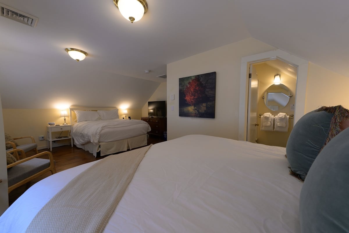 A hotel room with a large bed, bedside tables, a chair, a painting, and a bathroom with a mirror and towel rack.