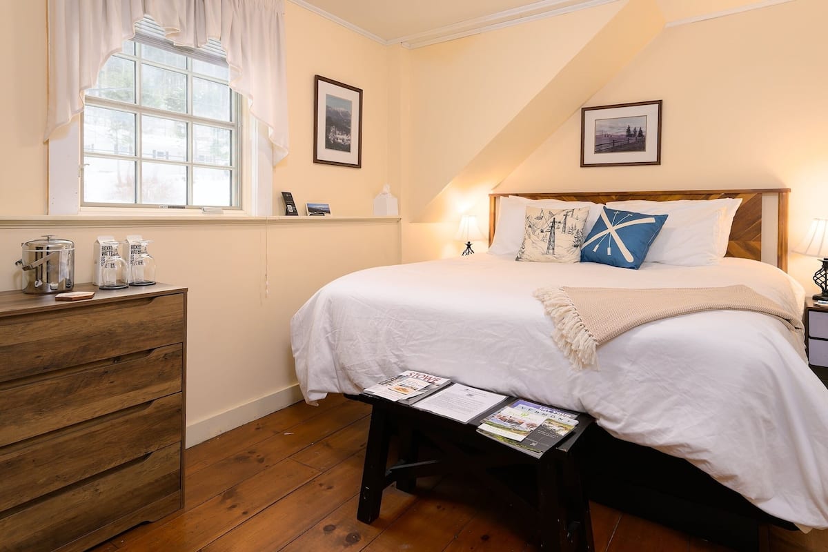 A bedroom with a large bed, a wooden dresser, a window with curtains, and a bedside table with a lamp and a magazine.