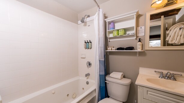 Un baño con bañera, inodoro y lavamanos. Hay estantes con artículos de tocador y un espejo sobre el lavamanos.