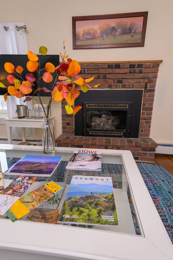 A living room with a brick fireplace, a vase with autumn leaves, and a glass table displaying travel brochures.