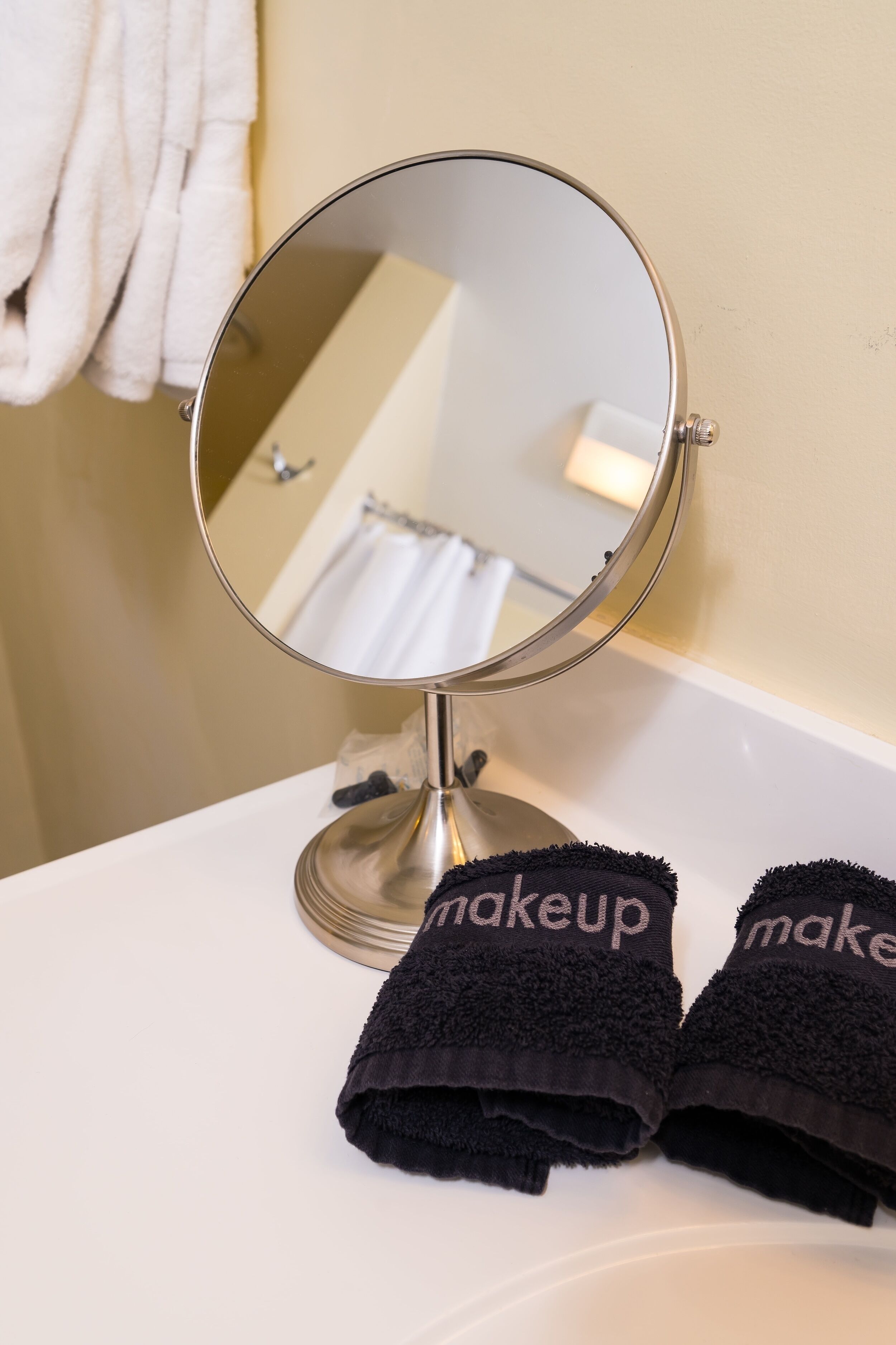 A round mirror on a white bathroom counter with two black towels labeled "makeup" beside it.