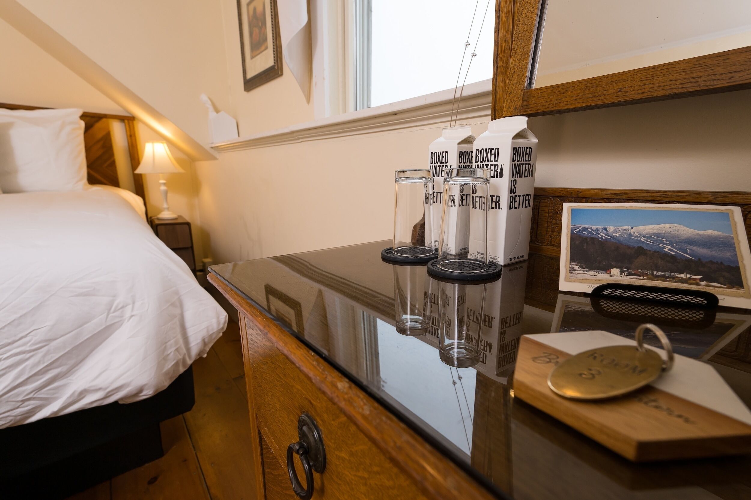 A hotel room with a bed, bedside table, and a desk with a computer monitor displaying a scenic image. There are three boxed water bottles on the desk.