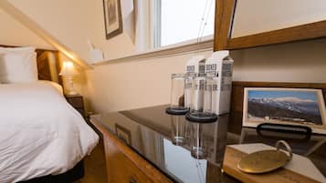 A hotel room with a bed, bedside table, and a desk with a computer monitor displaying a scenic image. There are three boxed water bottles on the desk.