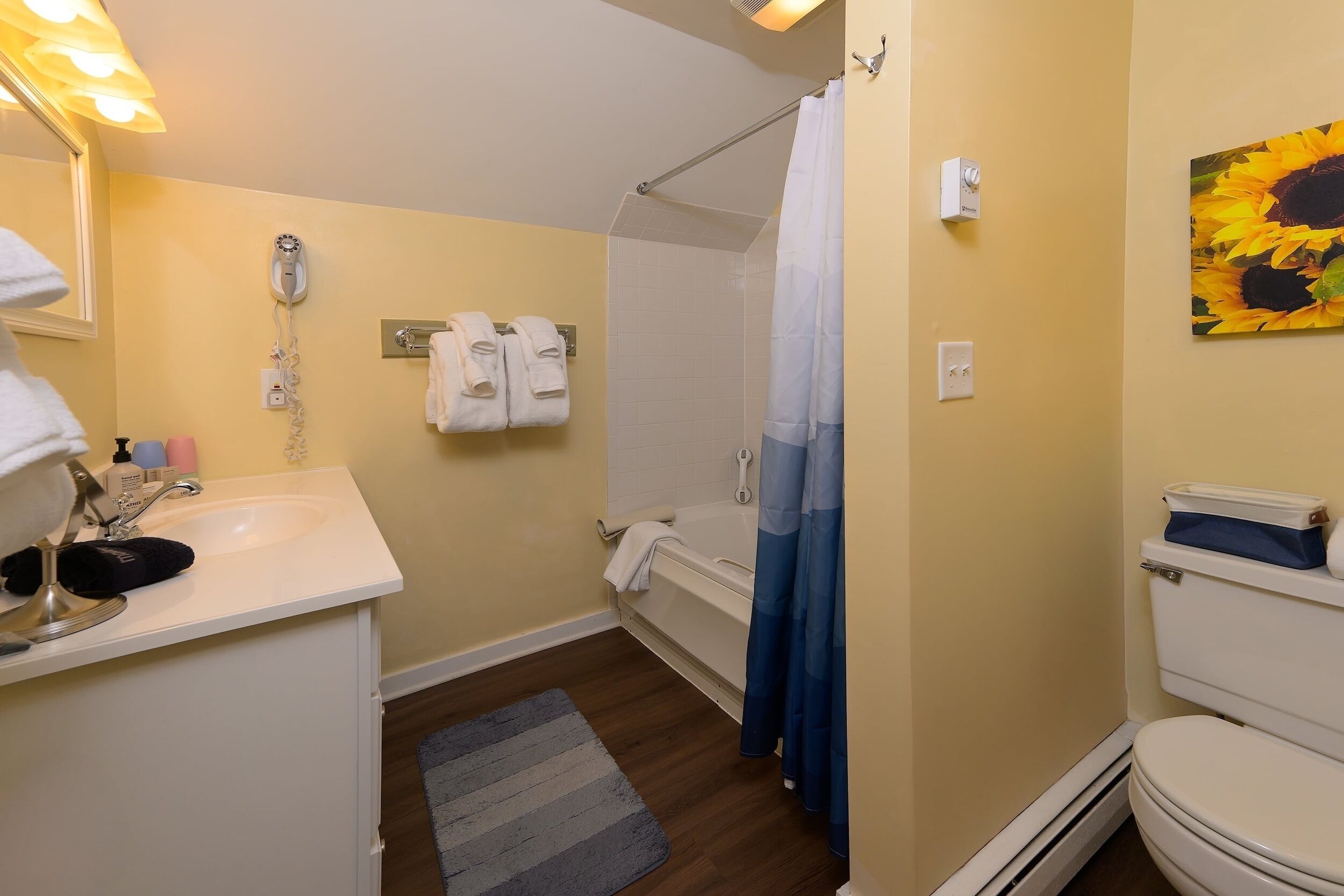 A bathroom with a sink, mirror, and a shower with a curtain. There are towels hanging on the wall and a painting of sunflowers.