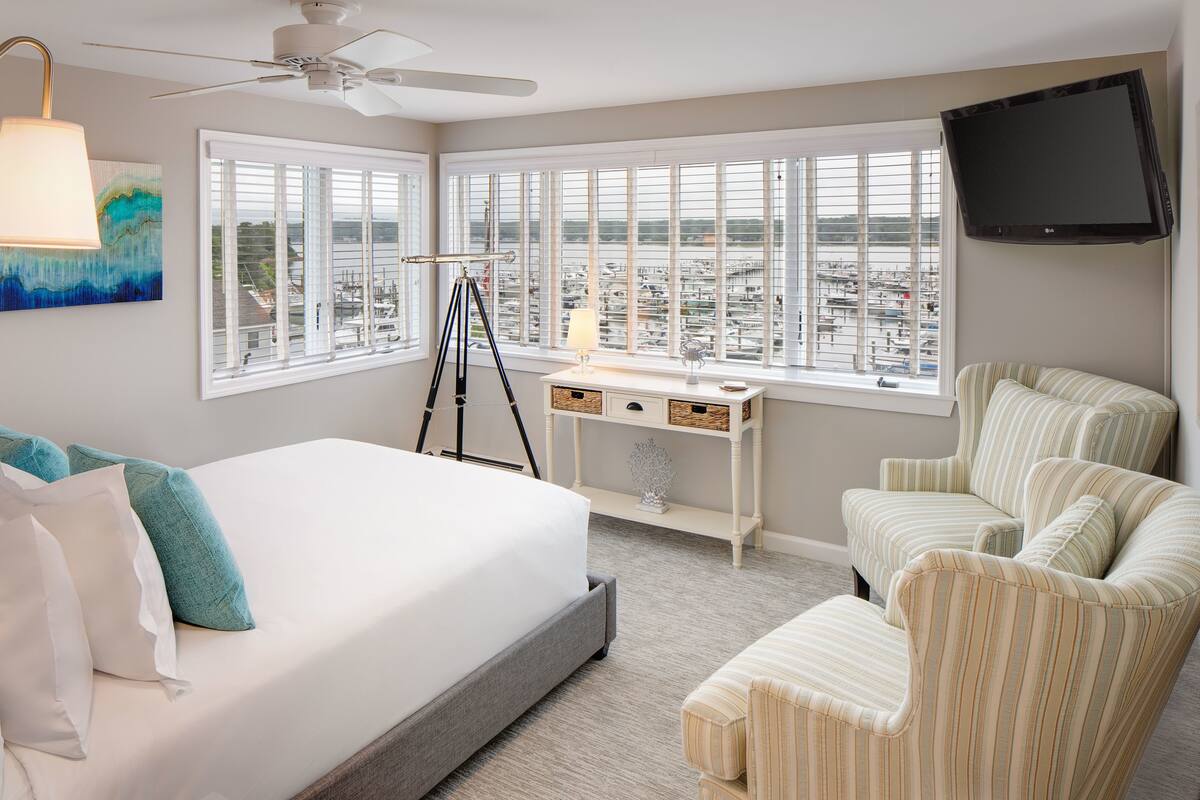 A hotel room with a bed, a desk, a chair, a television, and large windows offering a city view.
