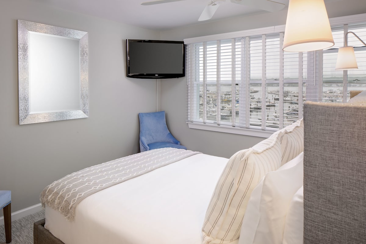 A hotel room with a bed, a television, a window with blinds, and a city view.