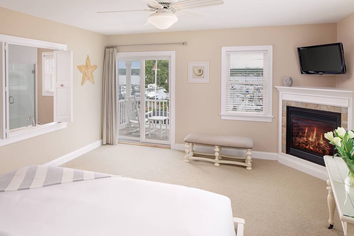 A bedroom with a large bed, a fireplace, a television, and a balcony with a view of boats.