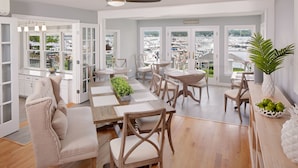Un comedor amplio con varias mesas y sillas, ventanas grandes con vista al puerto deportivo y una cocina moderna con armarios blancos.