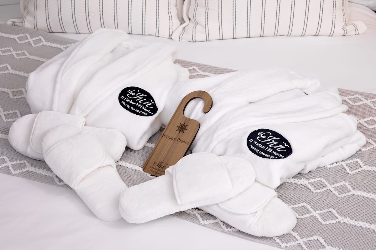 Two white bathrobes with wooden tags on a bed with striped pillows and a patterned blanket.