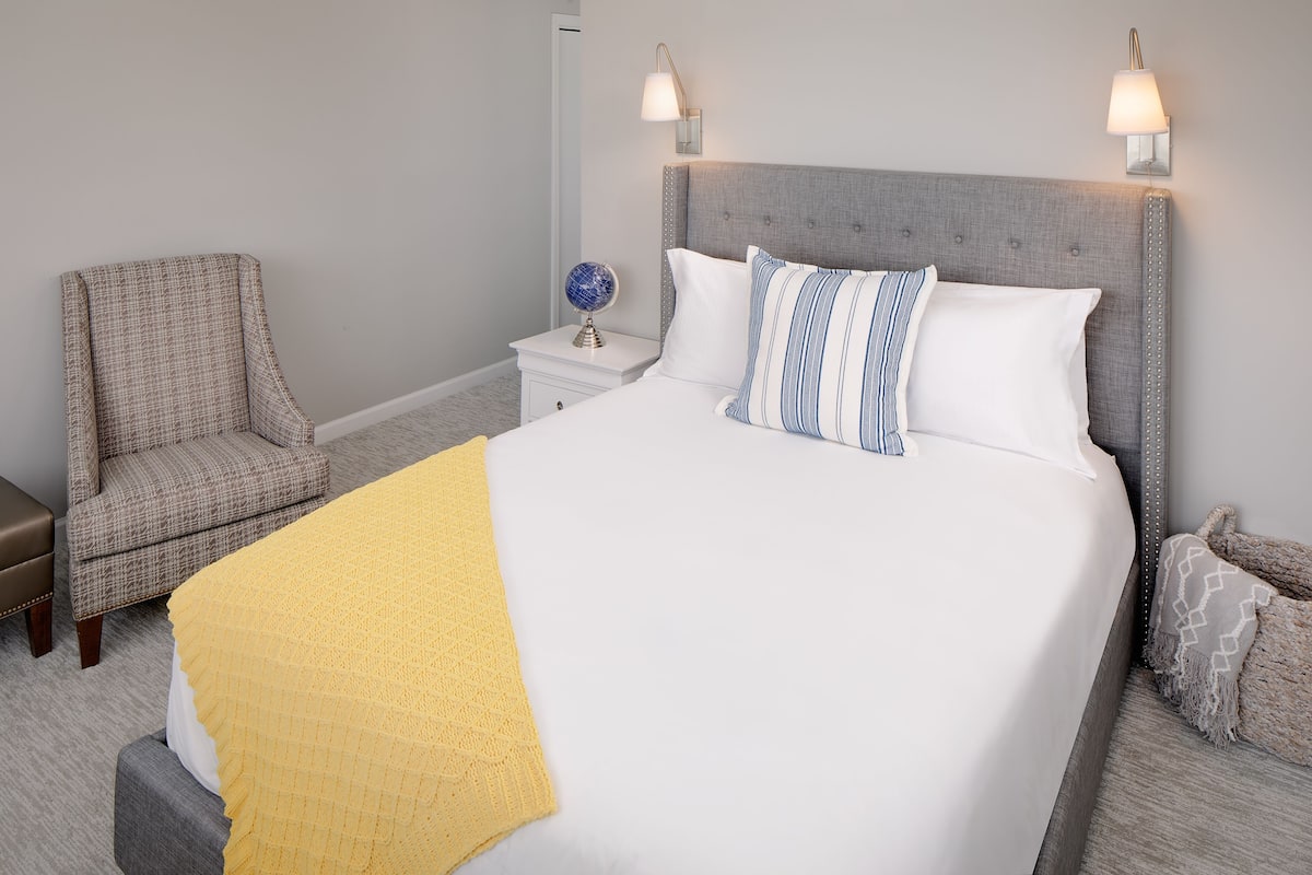 A neatly arranged bedroom with a large bed, a chair, a nightstand with a lamp, and a wall-mounted light fixture.
