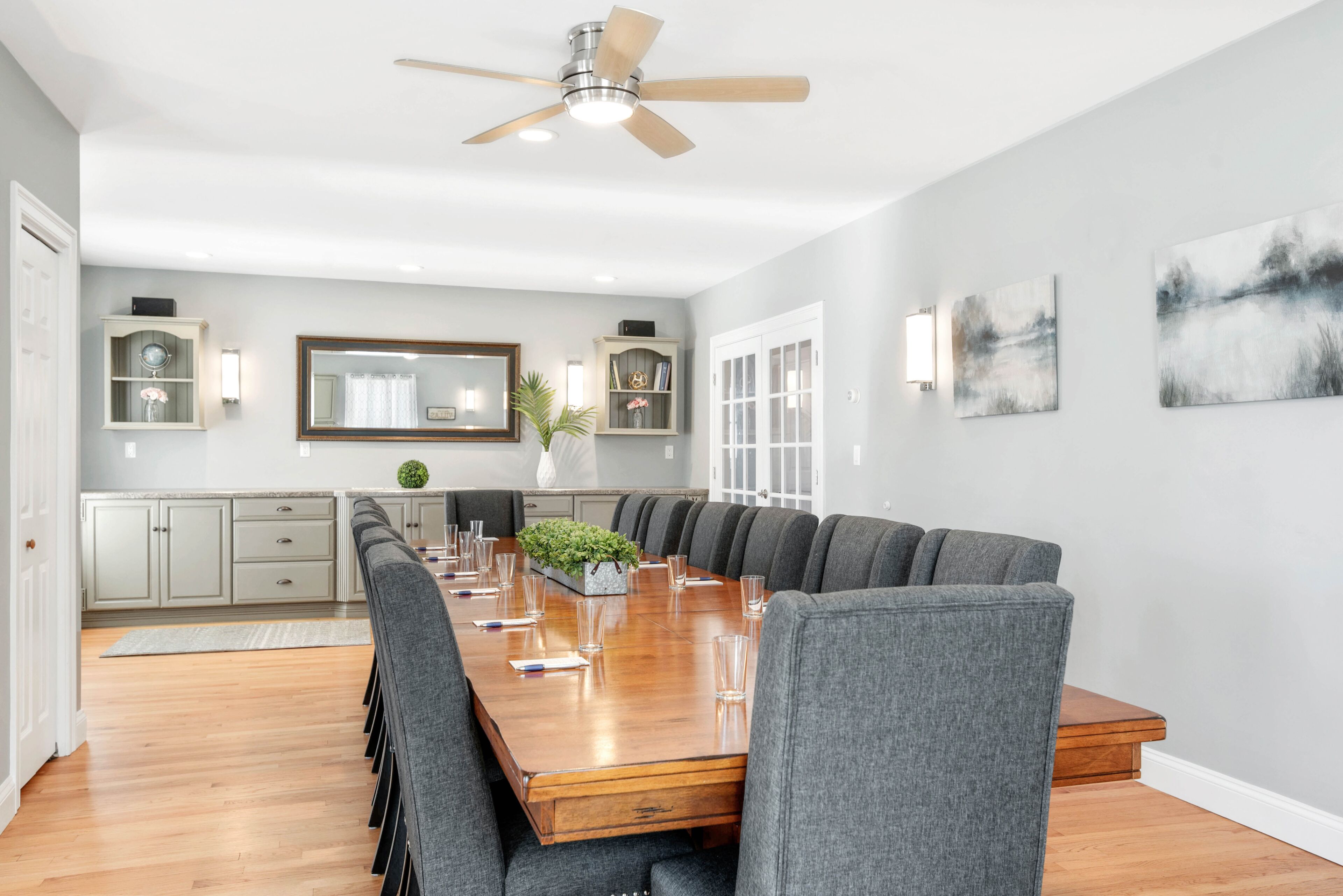 A modern dining area with a long wooden table, grey upholstered chairs, and a ceiling fan.