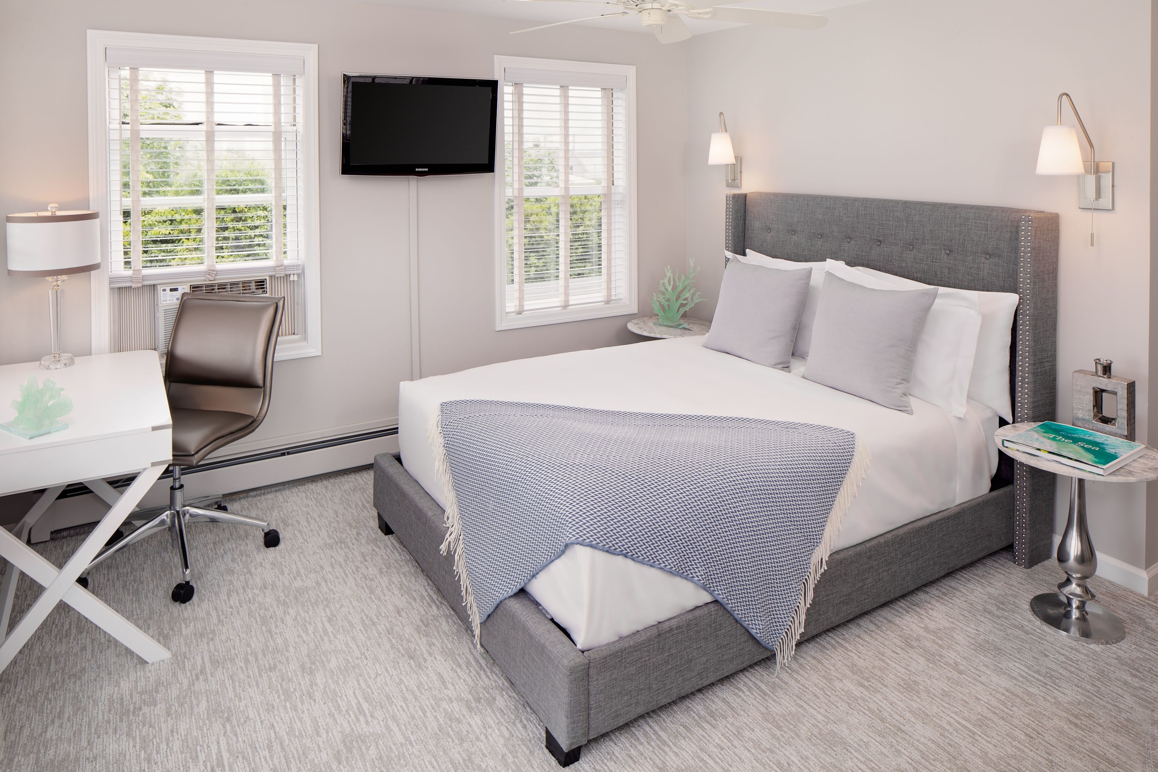 Habitación de hotel con cama, escritorio con silla, televisión montada en la pared y ventana con persianas.