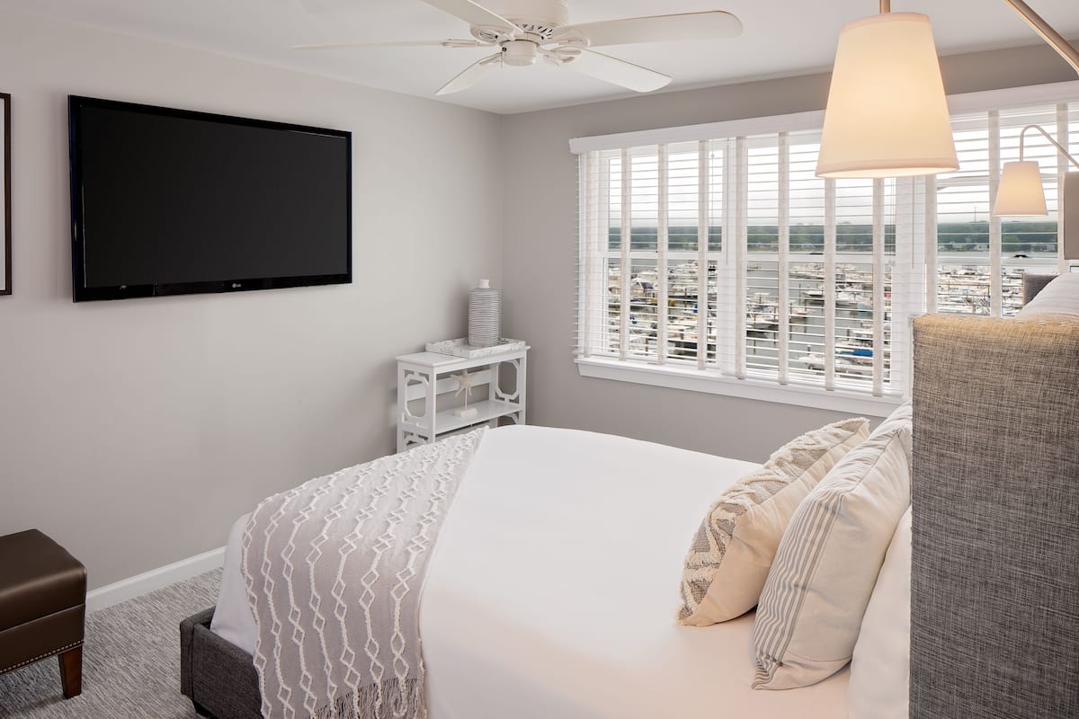 A bedroom with a bed, a TV, a ceiling fan, a window with blinds, and a view of a marina.