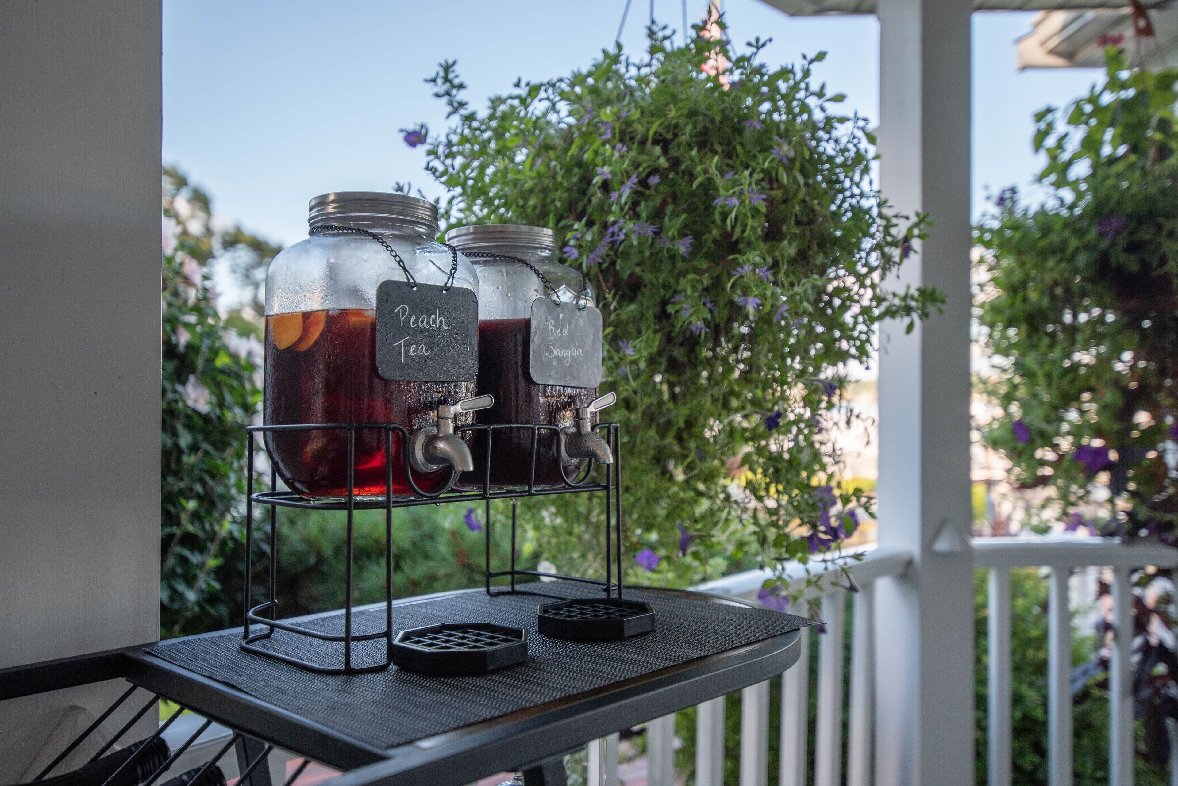 A beverage station with two labeled jars, one for peach tea and one for red sangria, on a balcony with a white railing and greenery in the background.