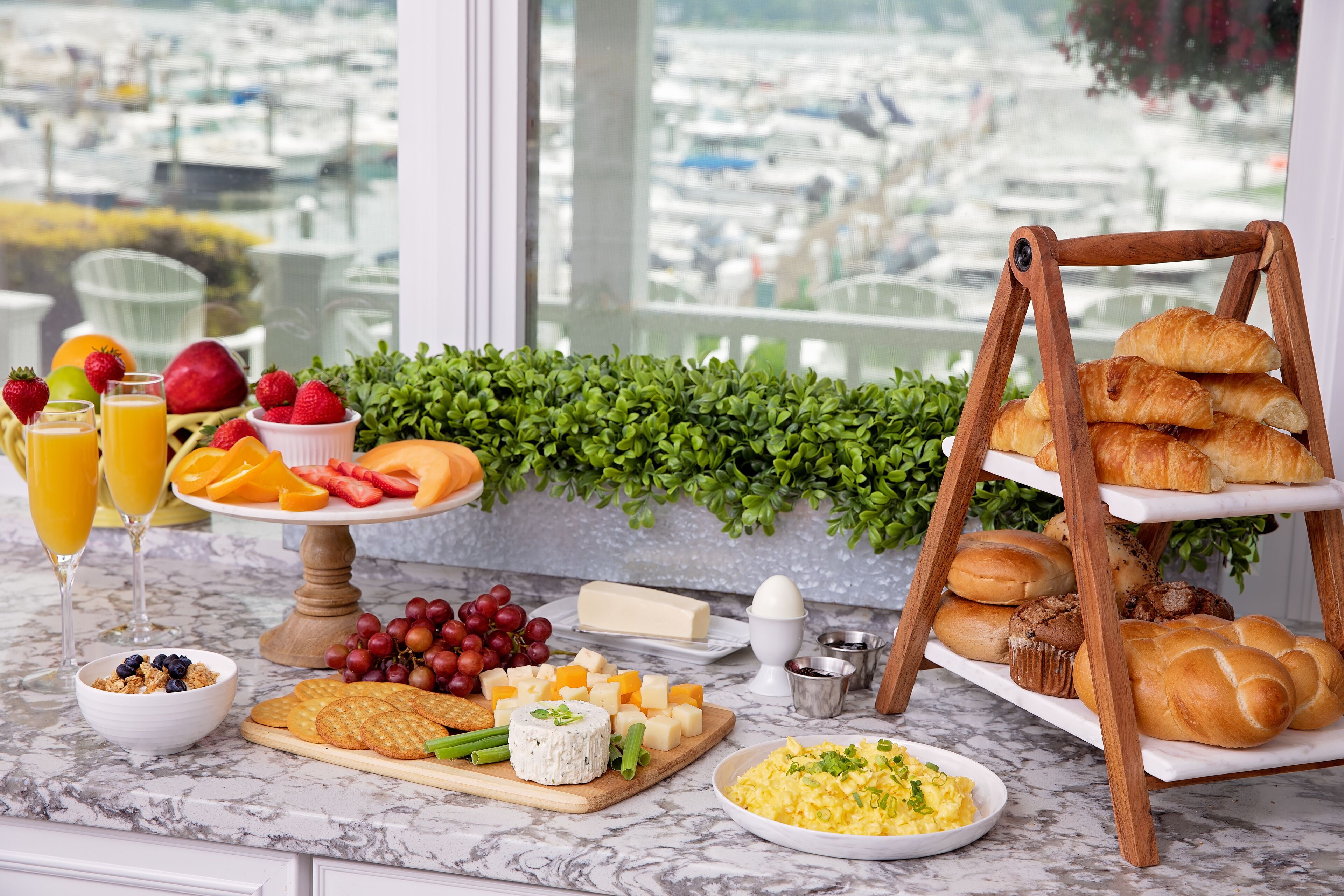 A breakfast buffet with various foods and drinks, including fruits, cheeses, pastries, and a view of boats through a window.
