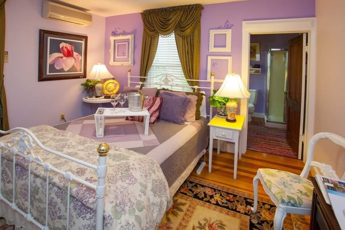 Un dormitorio con cama, mesitas de noche, lámparas, una silla y una puerta que da a otra habitación.