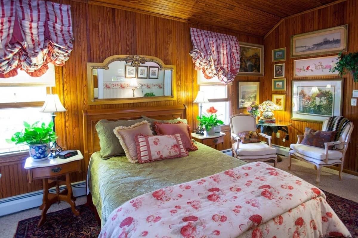 Un dormitorio acogedor con techo de madera, una cama grande con ropa de cama floral, dos mesitas de noche con lámparas, una silla y cuadros en las paredes.