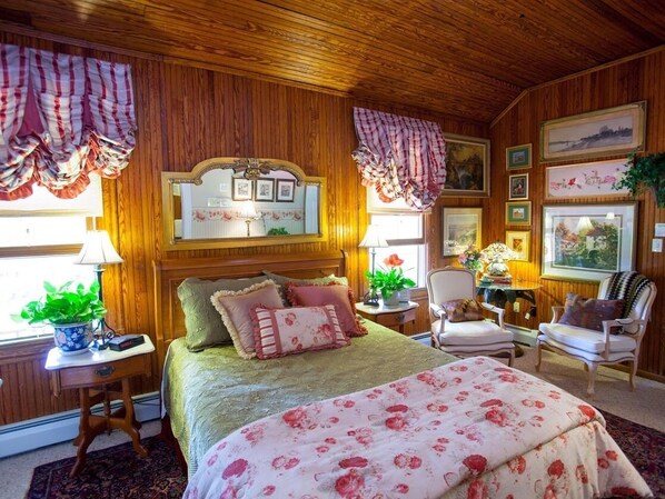 Un dormitorio acogedor con techo de madera, una cama grande con ropa de cama floral, dos mesitas de noche con lámparas, una silla y cuadros en las paredes.