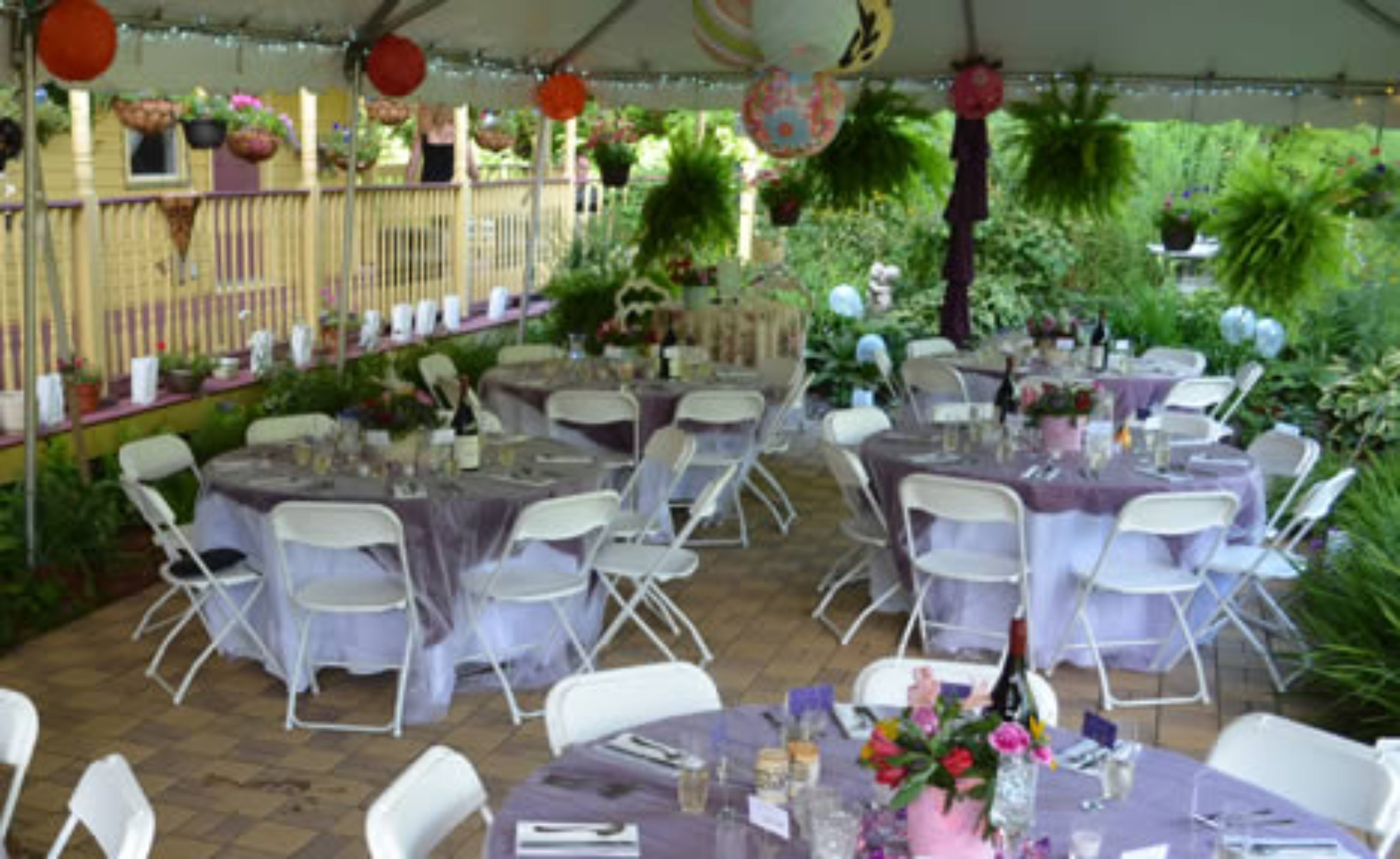 An outdoor event space with round tables covered in white tablecloths, surrounded by white chairs.
