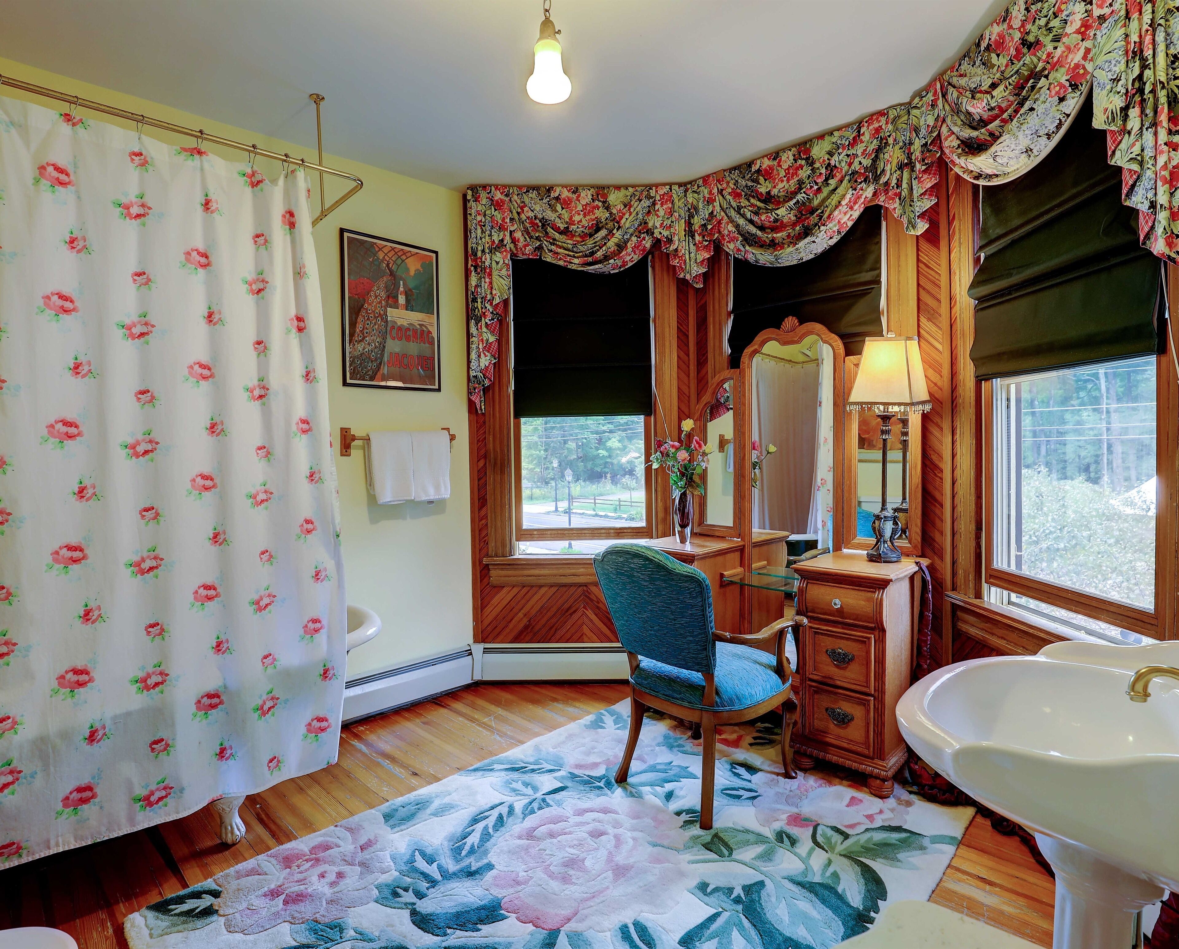 A bathroom with a wooden vanity, a sink, a chair, a floral rug, a shower curtain with a floral pattern, and a window with a view of greenery.
