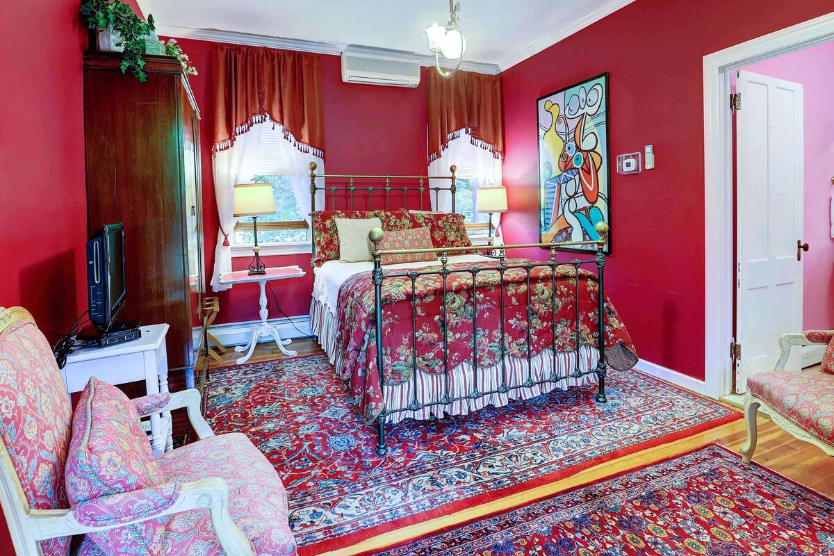 Un dormitorio con una pared roja, una cama con colcha estampada, una mesita de noche blanca, una lámpara y un cuadro grande.