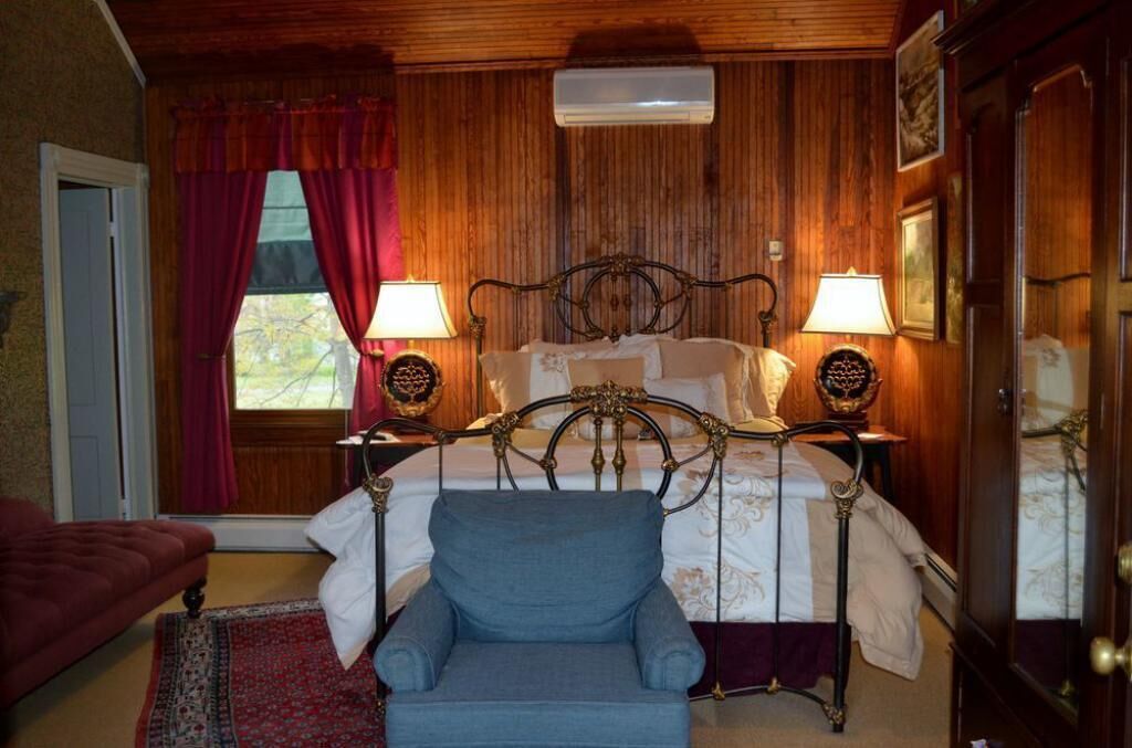 A bedroom with a wooden paneled wall, a bed with ornate headboard, two bedside lamps, and a blue armchair.
