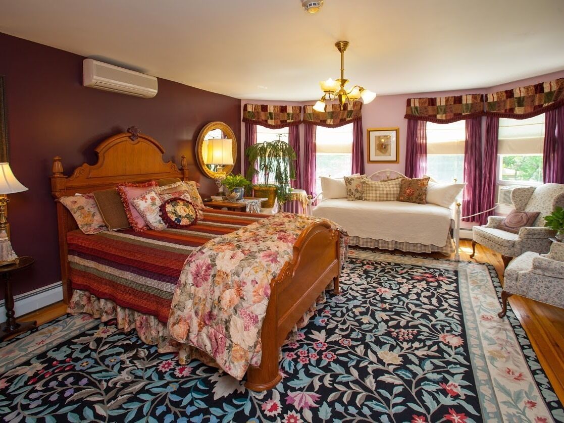 Un dormitorio con una cama grande, una cama más pequeña, una silla y una mesita de noche. Hay una ventana con cortinas, un candelabro y un espejo en la pared.