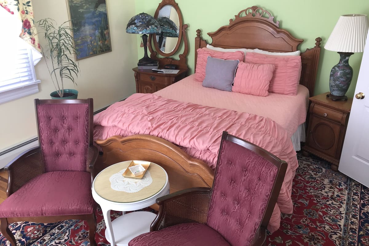 Un dormitorio con cama de madera, dos sillas, una mesita redonda, una mesita de noche con lámpara, un espejo y una planta en maceta.