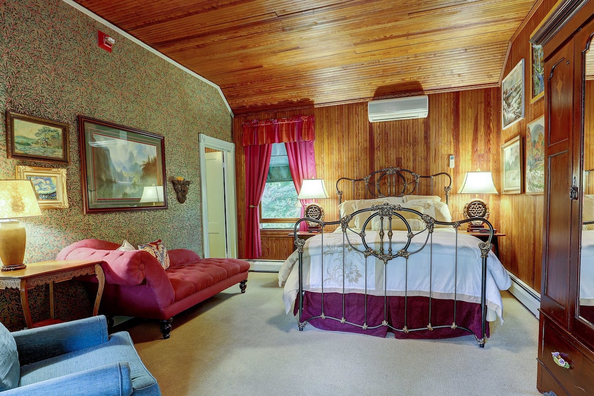 Un dormitorio con techo de madera, una cama con estructura metálica, un sofá rojo, un sillón azul y un armario de madera.
