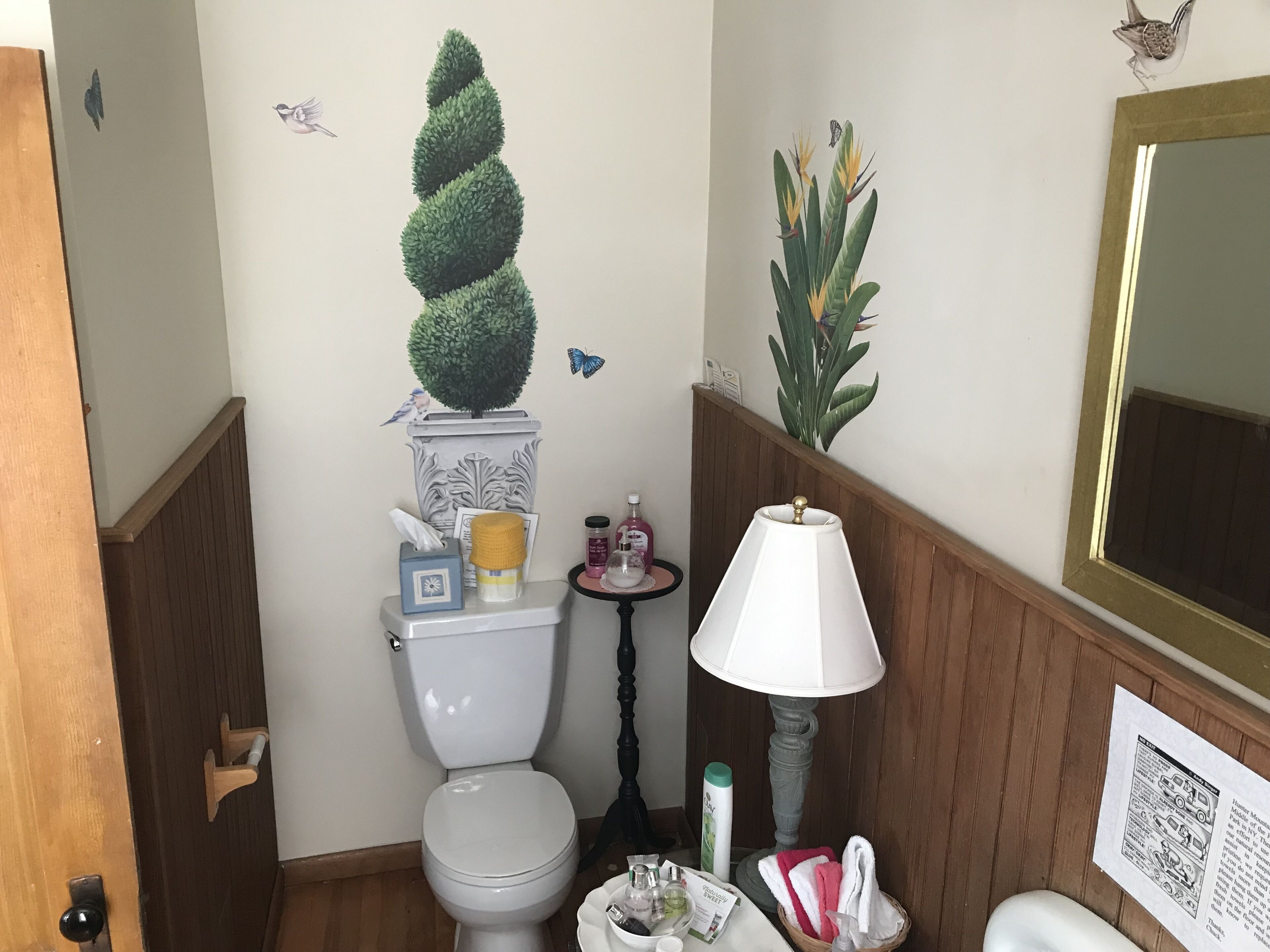 Un baño con inodoro, una mesita con artículos de tocador, una lámpara y decoración en la pared.