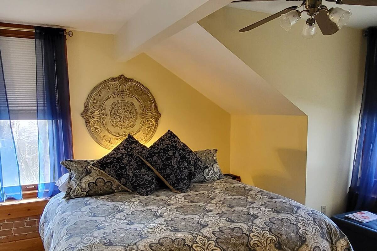 A bedroom with a large bed, a ceiling fan, a decorative wall piece, and a view of the outdoors through a window.