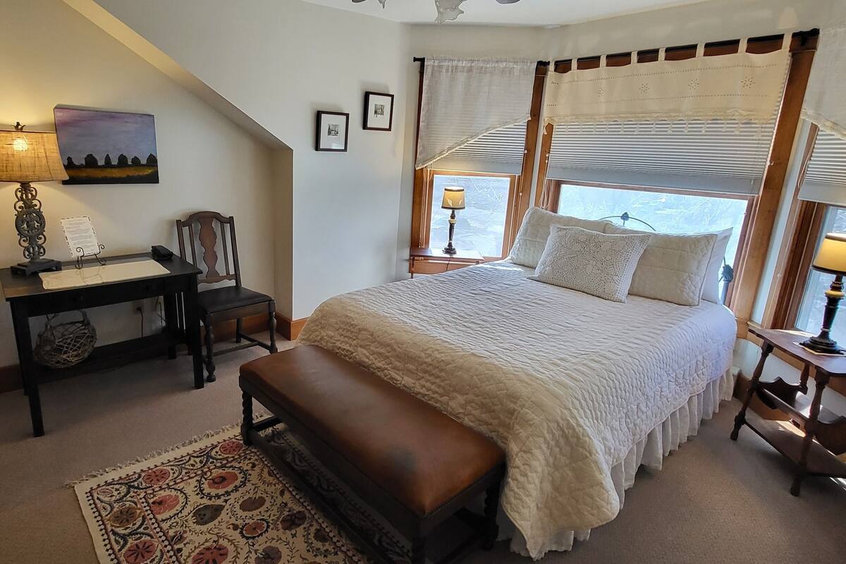 A bedroom with a bed, a desk, a chair, a bench, and a window with curtains.