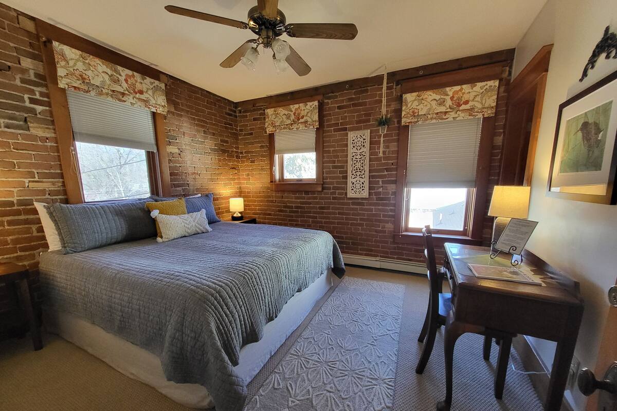 A bedroom with a brick wall, a bed with a grey quilt, a desk with a lamp, and a ceiling fan.