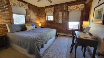 A bedroom with a brick wall, a bed with a grey quilt, a desk with a lamp, and a ceiling fan.