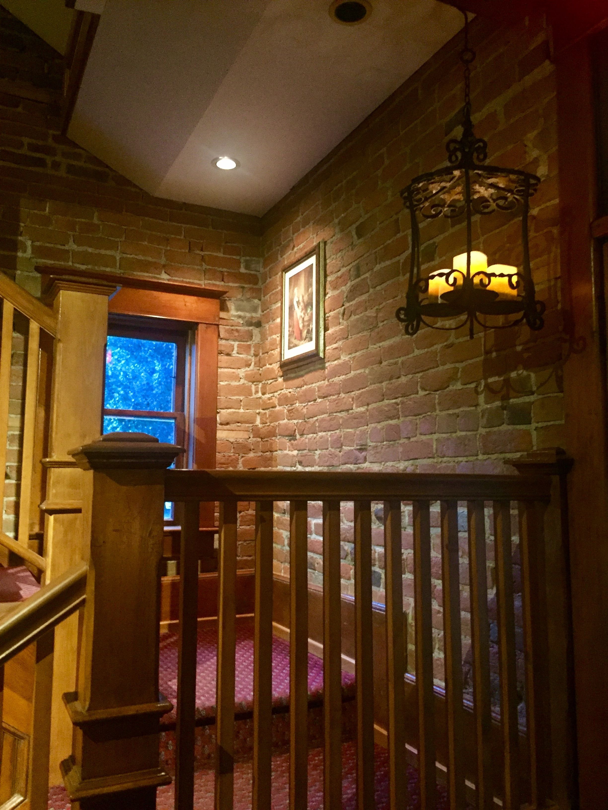 A staircase with wooden railings leading to a room with a brick wall, a framed picture, and a hanging light fixture.