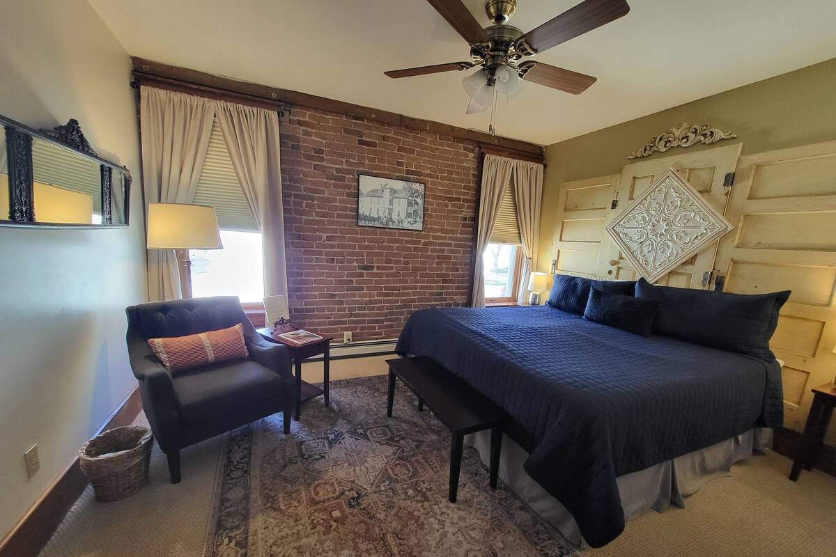 A bedroom with a large bed, a chair, a small table, a ceiling fan, and a brick wall.