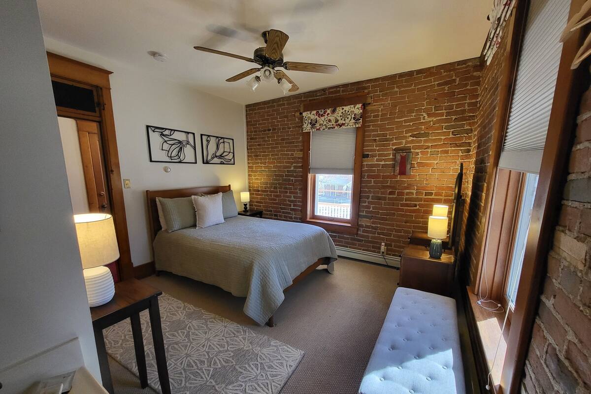 A bedroom with a bed, two bedside lamps, a ceiling fan, a brick wall, and a window with blinds.