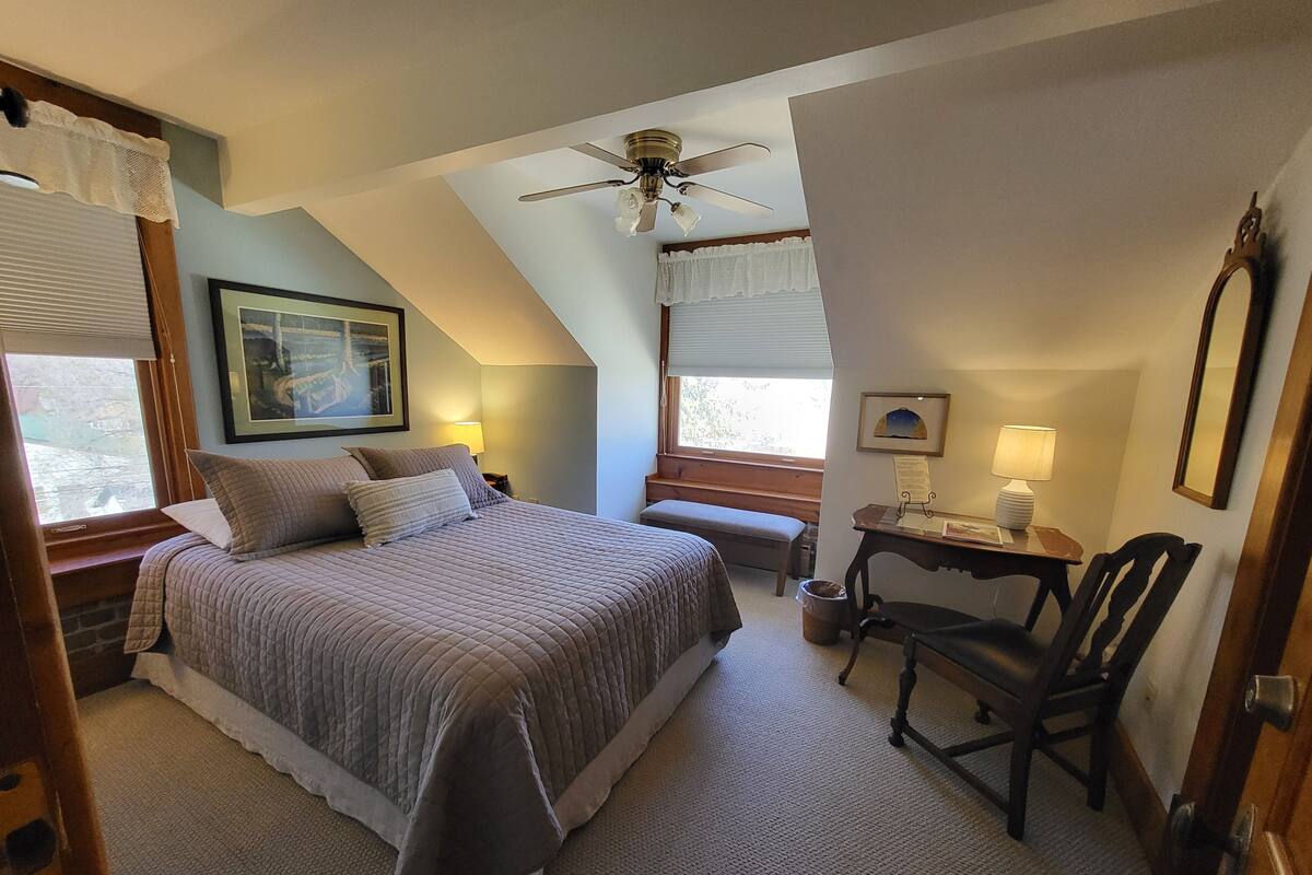 A bedroom with a bed, a desk, a chair, a ceiling fan, and a window with blinds.
