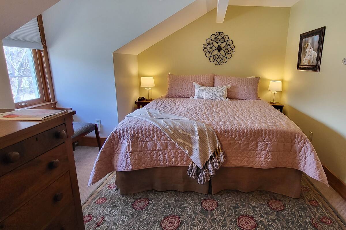 A bedroom with a large bed, two bedside lamps, a wooden dresser, a patterned rug, and a decorative wall piece.