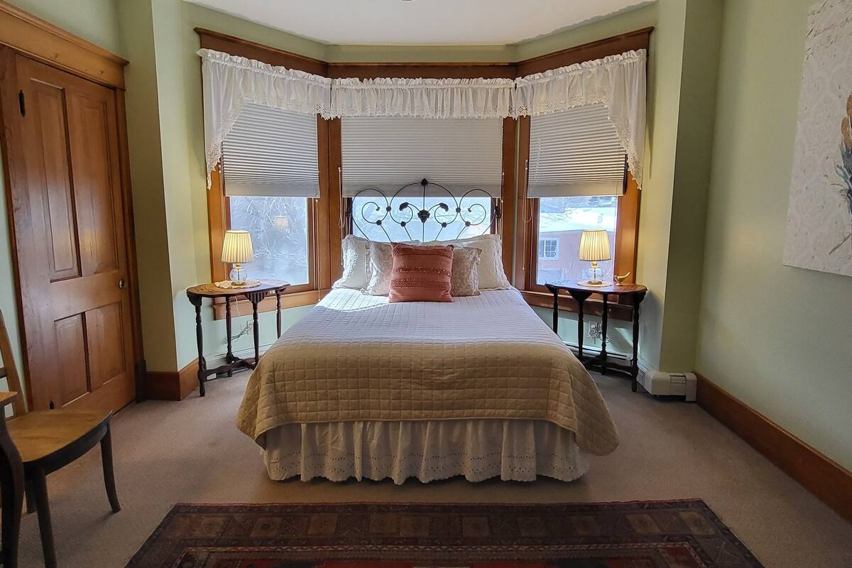 A bedroom with a bed, two bedside tables with lamps, a chair, a wooden door, and a framed artwork on the wall.