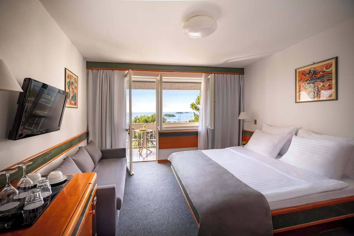 Quarto de hotel com cama, televisão, escrivaninha com bandeja de copos, janela com cortinas e um quadro na parede.