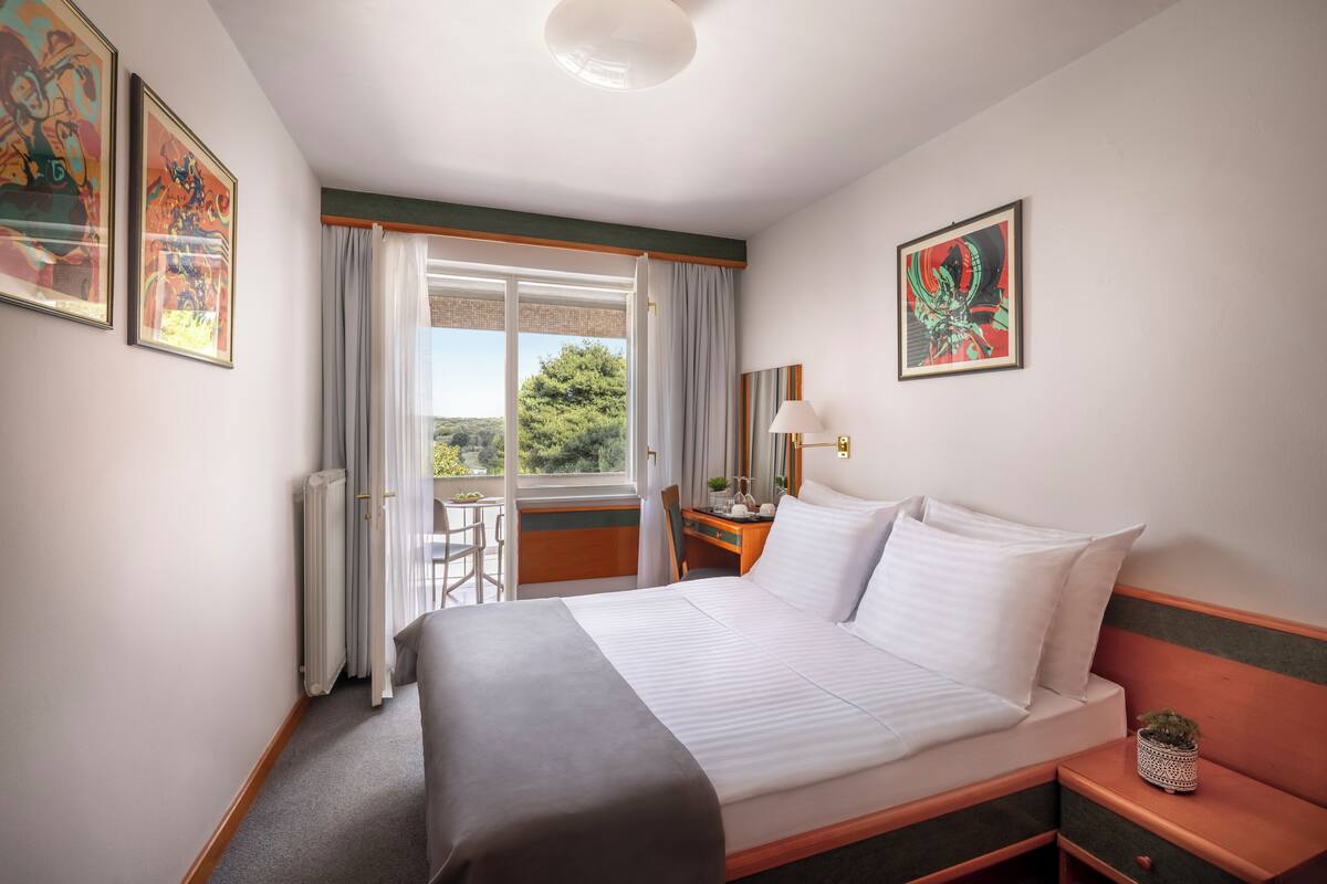 Quarto de hotel com cama, escrivaninha, cadeira, luminária e vista para o exterior.