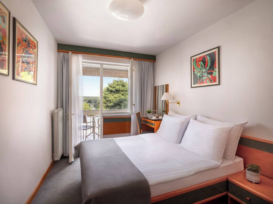 Habitación de hotel con cama, escritorio, silla, ventana con cortinas y obras de arte en las paredes.