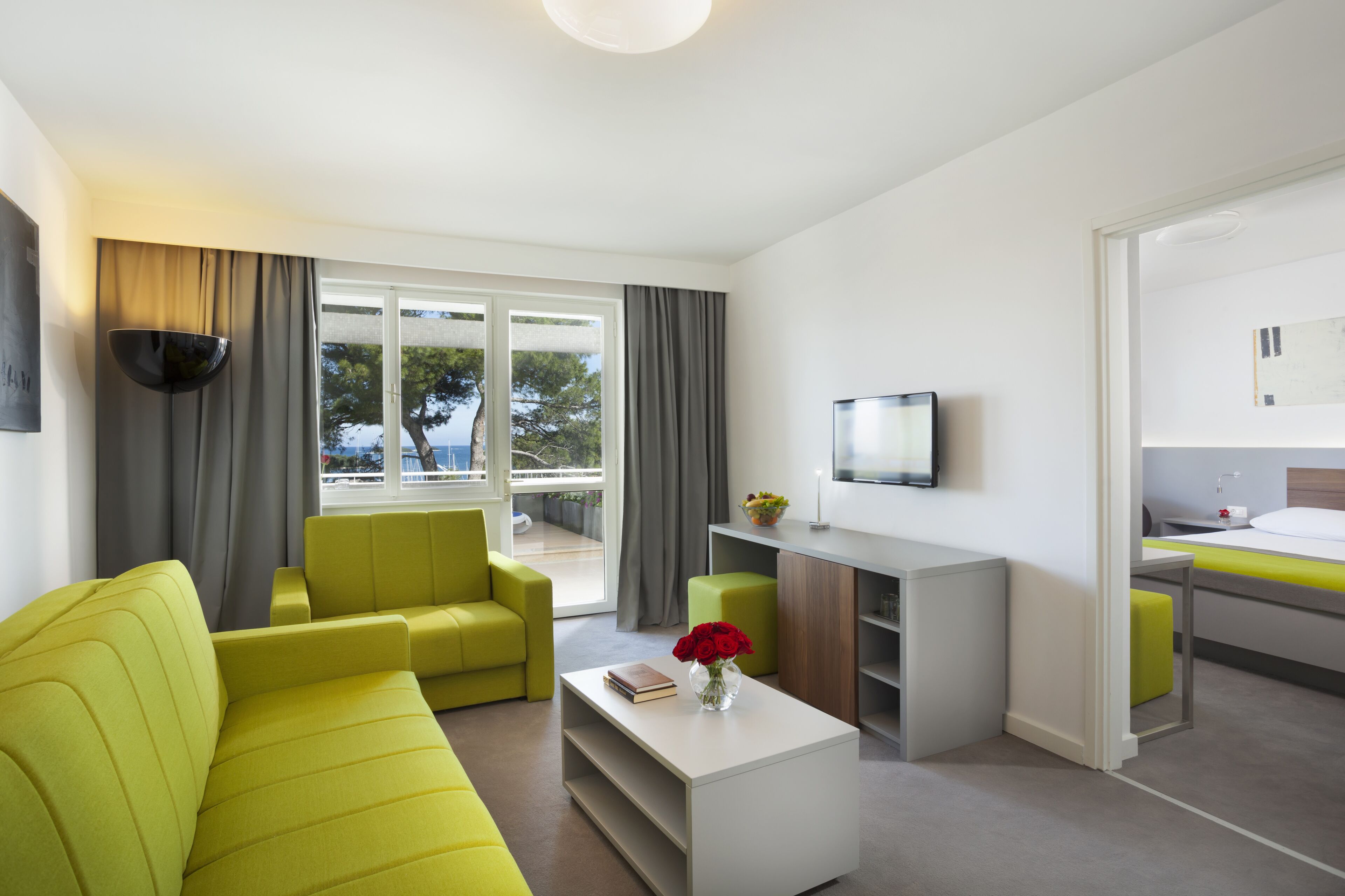 A modern hotel room with a sofa, a coffee table, a TV, a small desk, and a view of the sea through large windows.