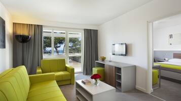 A modern hotel room with a sofa, a coffee table, a TV, a small desk, and a view of the sea through large windows.