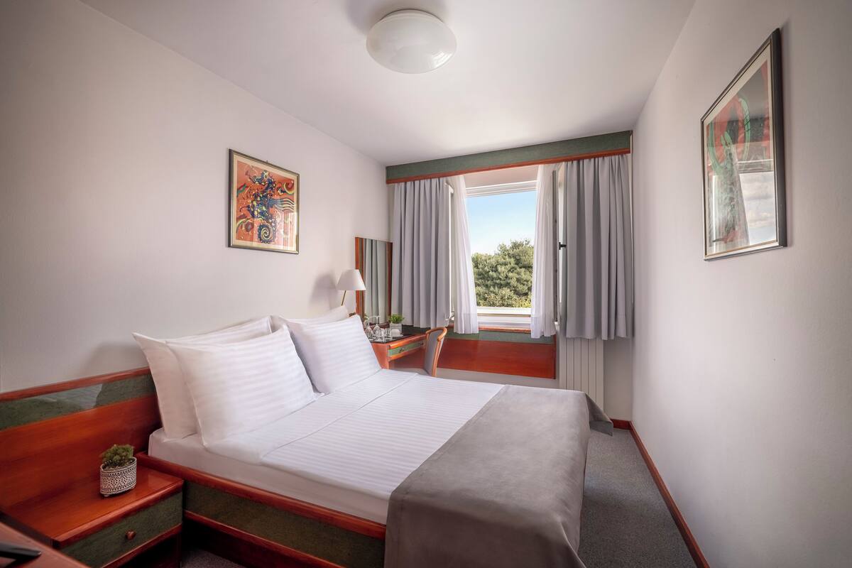 Quarto de hotel com cama, mesa de cabeceira, luminária e janela com cortinas.