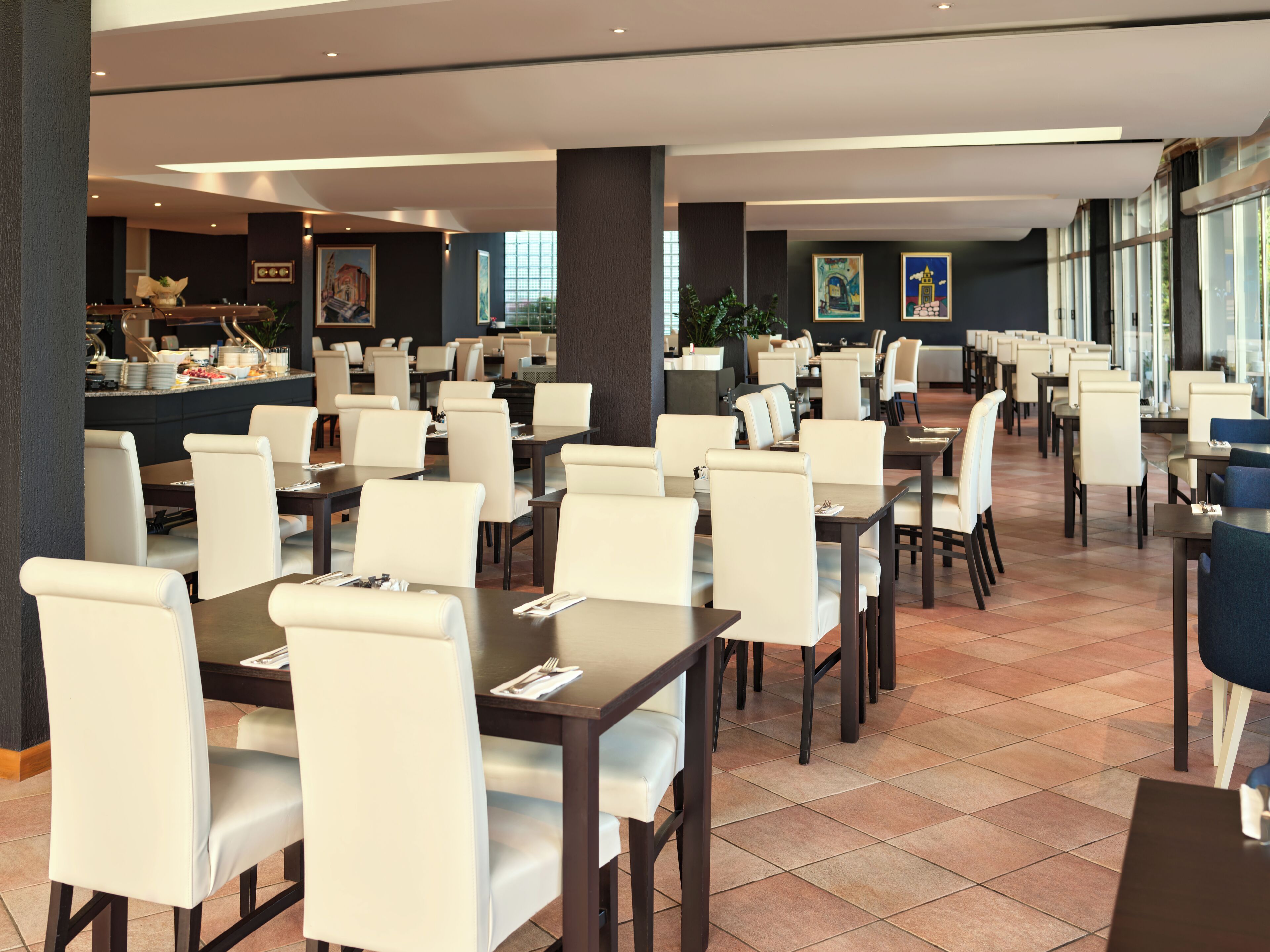 A spacious dining area with multiple tables and chairs, a buffet setup, and large windows providing natural light.