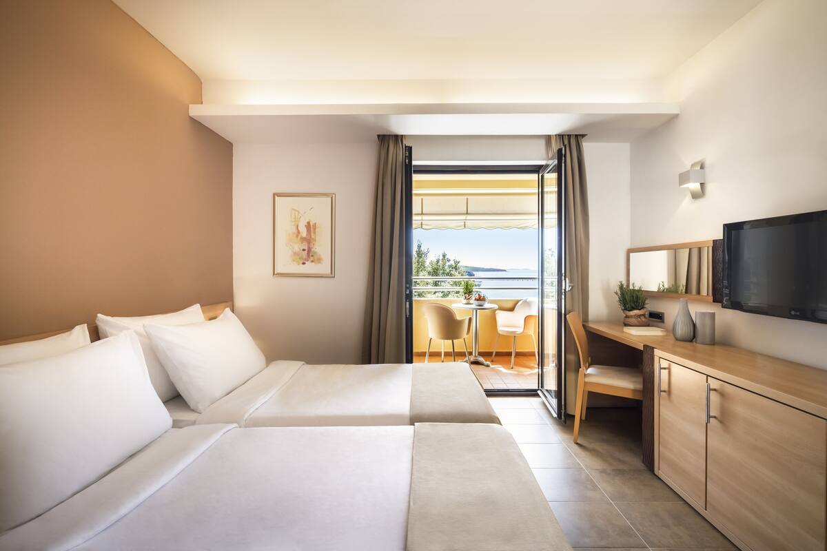 A hotel room with two beds, a desk with a TV, and a balcony with a view.