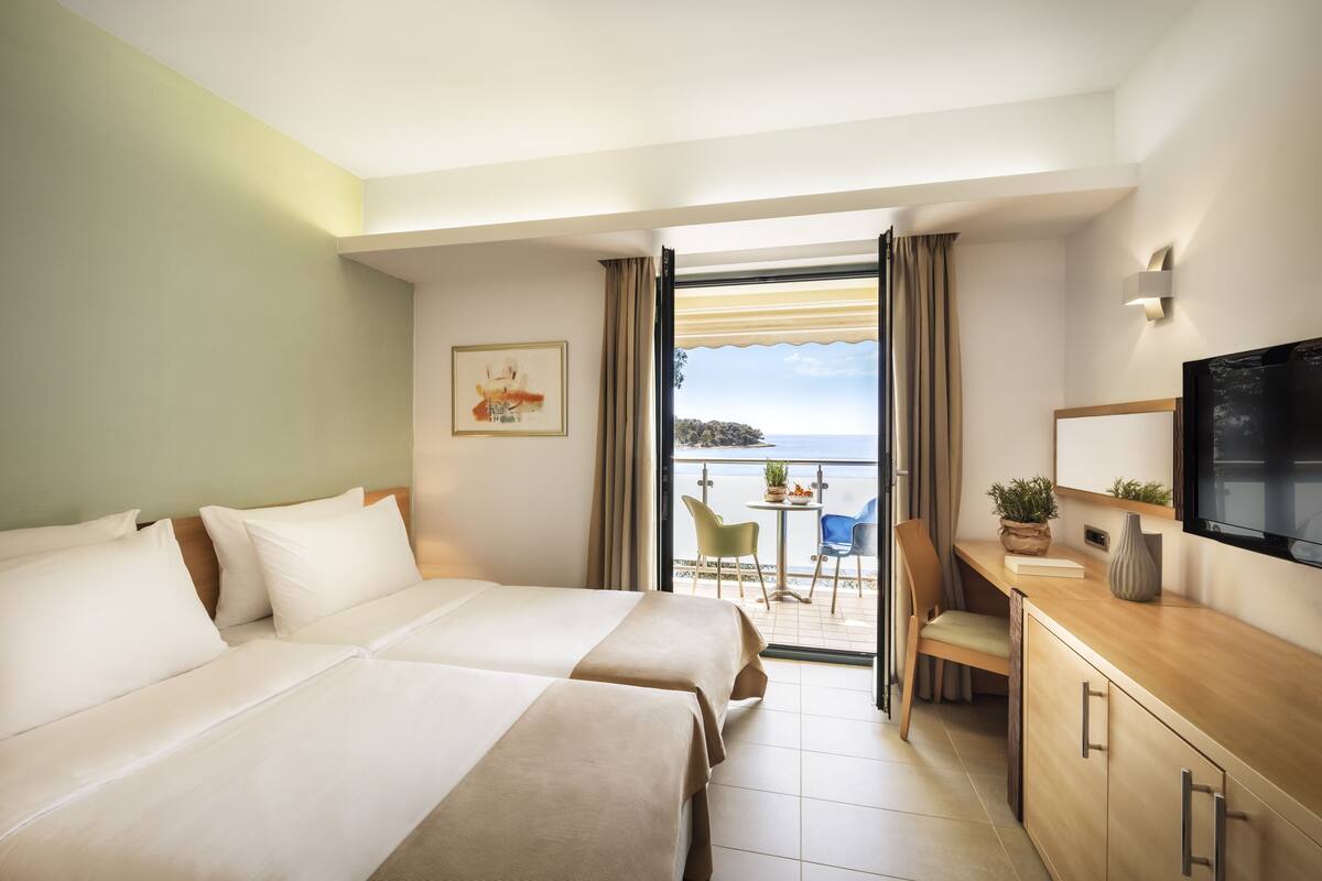 A hotel room with two beds, a desk, a TV, and a balcony with a view of the sea.