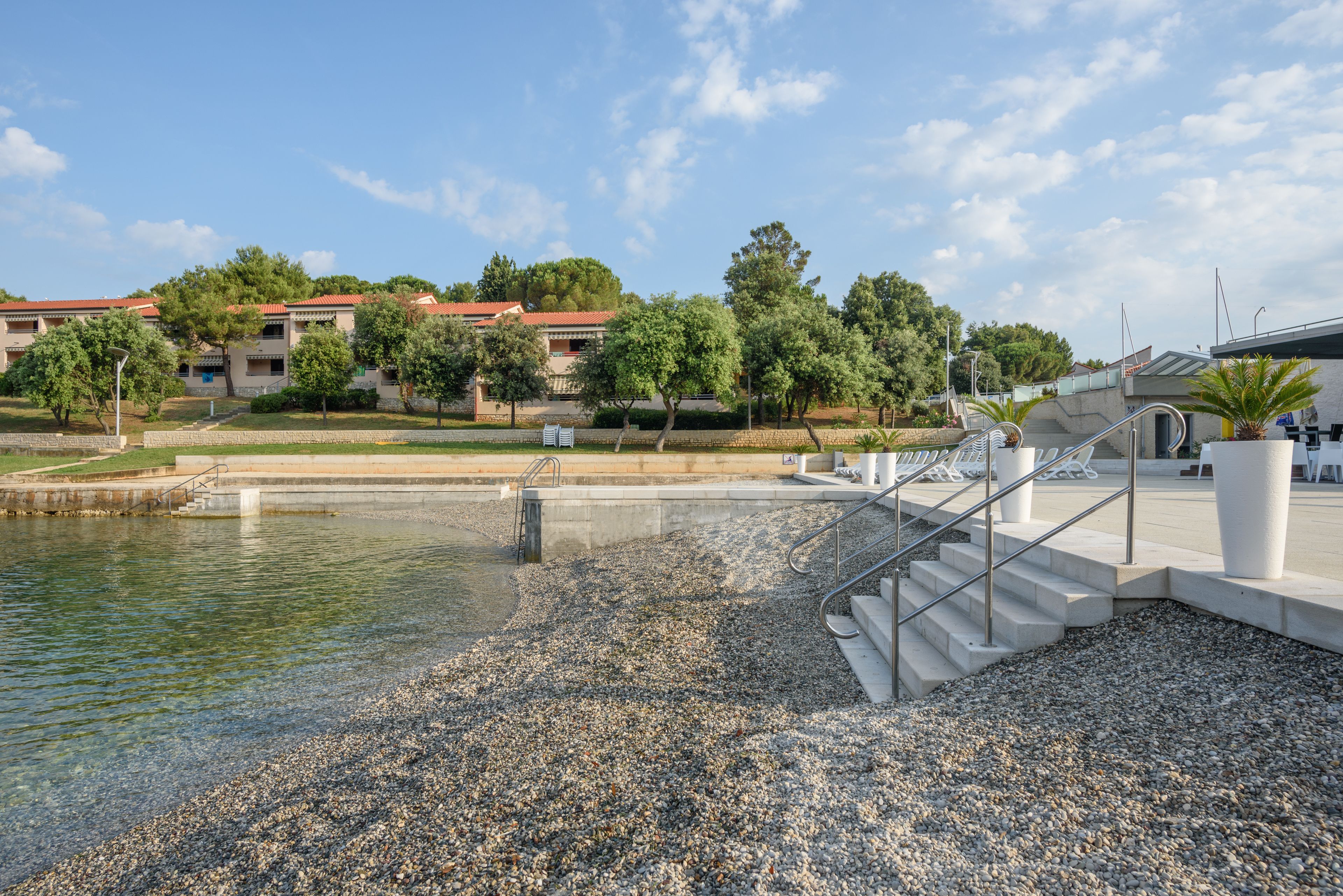 Un hôtel situé au bord de l’eau, avec une plage de galets, un escalier menant à une piscine et, en arrière-plan, un bâtiment aux toits rouges.